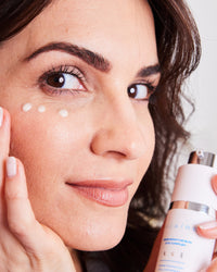 Close-up of woman applying white cream near eye holding Triple Threat Trio bottle gallery position 1